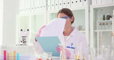 Gözlüklü kadın bilim adamı laboratuardaki dokümanları inceledi. Kadın kimyager, test tüpleri ve mikroskopla masada rapor okuyor.