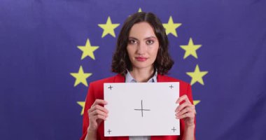 Politikacı elinde üzerinde artı işaretleri olan kağıtlar tutuyor. Artı işaretler, kadınların AB bayrağına karşı büyümeyle ilgili konuşmalarını güçlendiren güçlü bir görsel yardım görevi görüyor