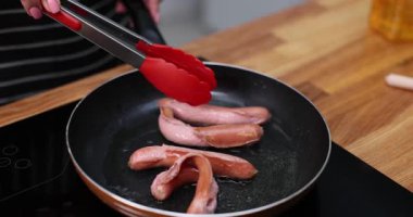 Kadın, mutfakta maşayla sıcak tavada lezzetli sosislere dönüşüyor. Bayan, indüksiyon sobasında et malzemeleriyle besleyici bir kahvaltı hazırlıyor.