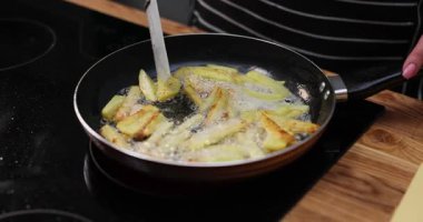 Kadın, indüksiyon sobasında patates dilimlerini kaynayan bitki yağında kızartıyor. Önlüklü bir kadın, aydınlık mutfakta akşam yemeği için yağlı ve sağlıksız yemekler pişirir.