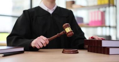 Siyah cübbe giyen bir yargıç, mahkeme salonu dolusu kitap ve belgelerle dolu bir tokmak tutuyor.