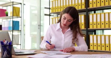 Genç bir kadın, görevlerine odaklanmış, parlak ve düzenli bir ofis alanında çeşitli belgelerle özenle çalışıyor.