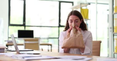 Masada oturan genç bir kadın, kahverengi bir kesekağıdının içine nefes alıp veriyor, işyerinde stres ya da kaygı belirtileri gösteriyor..