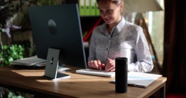 Bir kadın şık bir ofiste çalışıyor, bilgisayarında akıllı bir hoparlörün yanında yazarak verimliliğini arttırıyor.