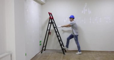 Çalışkan bir işçi, sağlam bir merdiven kullanırken ve gerekli güvenlik malzemelerini giyerken iç duvarı boyamaya hazırlanıyor.