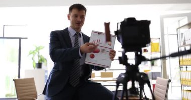 Bir iş adamı, şık bir ofis ortamında bir belgeyi sunarken seyirciyle ilişki kurar..