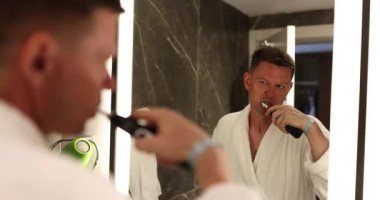 Rahat bir bornoz giymiş bir adam, zarif bir banyo aynasının önünde dişlerini fırçalar, harmanlama tarzı ve konforu vardır.