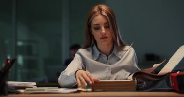 Kadın ofis çalışanı, evraklarla dolu büyük klasörlerin sayfalarını inceliyor. Kadın çalışmaları muazzam bir klasörde depolanmış. Yavaş çekim