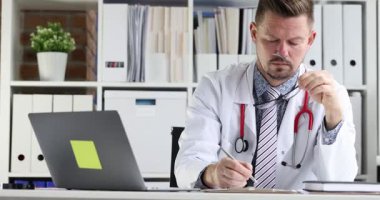 Odaklanmış sakallı doktor masada dizüstü bilgisayarla oturan belgeleri dolduruyor. Steteskoplu beyaz önlüklü adam klinikteki iş yerinde özenle çalışıyor.