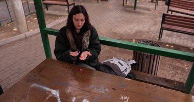 Genç kadın, ahşap masada kremalı nemlendirici ellere odaklanmış olarak oturuyor. Şehir parkında sakin bir kişisel bakım ve tefekkür anı