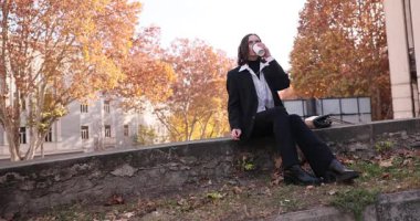 Kadın tesadüfen taş duvarda oturur sigara içmek ve kahve içmek için zaman ayırır. Sonbahar yapraklarıyla ağaçların yanında gevşemiş insan, dingin olduğunu gösteriyor.