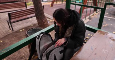 Genç bayan masada oturuyor ve sırt çantasının içindekilerle etkileşime geçiyor. Kişi, soğuk sonbahar havasında çantanın içindeki eşyaları aramaya ve düzenlemeye odaklanır.