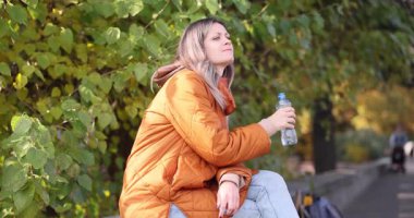 Genç bayan pervazda oturup plastik su şişesinden su içiyor. Yeşil yapraklı ağaçların yanında dinlenen insan, sıcak bir günde doğal park kurulduğunu öne sürüyor.