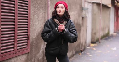 Kırmızı bereli ve koyu renk ceketli bir kadın dar bir sokakta yıpranmış duvarlarla dikilirken sigara içiyor. Şehrin daha eski kesimlerinde kentsel ortam