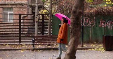 Parlak pembe şemsiyeli bir kadın sonbahar günü eski kasaba bahçesinde yürür. Turuncu ceketli kadın, şehirde ağır çekimde sakin bir gezinti yapıyor.