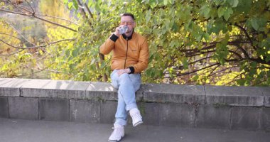 Deri ceketli adam su içiyor. Alçak kayalıklarda oturuyor. Erkek, şehir parkında sakin bir sonbahar gününün tadını çıkarıyor.