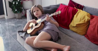 Küçük kız elinde gitarla koltukta rahatça uzanıyor. Yetenekli çocuk evde ukulele ile yavaş ve melodik bir melodi çalar. Yavaş çekim
