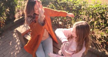 Genç kadın çocuğa beşlik çakıyor ve parkta bankta otururken sarılıyor. Güneşli bir günde anne ve kız arasındaki etkileşim