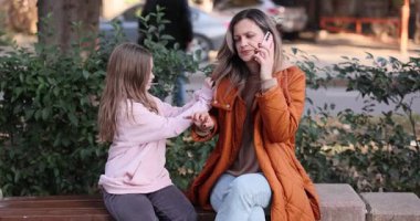 Kadın telefonu kulağına dayadı ve bankta oturarak konuşmaya başladı. Küçük kız dikkat çekmek için ısrarla annesinin kolunu çekiştiriyor.