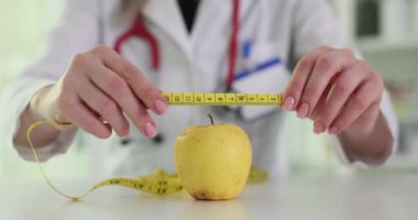 Palto giyen bayan doktor klinikte masasında parlak mezuralı elmanın ölçüsünü ölçer. Diyet ve porsiyon kontrolünün hayatta sağlık bağlamında önemi