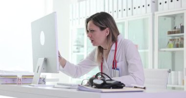 Açık renk ceketli kadın profesyonel, elinde kağıt tutarken dikkatle bilgisayar ekranına bakıyor. Tıp bürosu ortamında araştırma yapılıyor