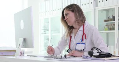 Kadın doktor bilgisayar ekranına odaklanıyor ve masada notlar alıyor. Ayarlar, hastanedeki hasta geçmişinin incelendiği sağlık ortamını öneriyor