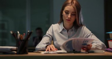 Genç bir kadın telefon başında dosyalarını gözden geçiriyor. İşçi ofisteki loş bir ortamda odaklanmış ve geç saatlere kadar çalışıyor gibi görünüyor