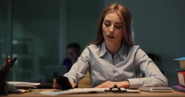 Telefonu masaya düşmüş bir kadın ofis ortamında yorgun ve stresli görünüyor. Akşamları geç ve fazla mesai ortamı vurgulanıyor
