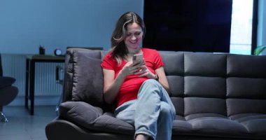 Gülümseyen kadın internette akıllı telefondan sohbet ediyor çağdaş kanepede dinleniyor. Cihaz mesajlı sarışın kadın cep telefonuyla kanepede oturuyor.