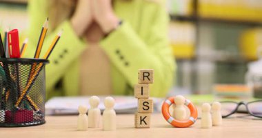Kadın masasında oturuyor, elleri, figürlerin ve can simidinin yanındaki bloklarda risk altında görünüyor. Ofis ortamında zorlu bir durum