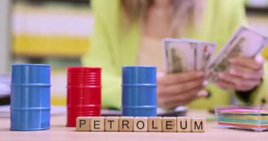 Ahşap bloklardaki petrol varillerinin masasında renkli petrol varilleri var. Sarı ceketli kadın işçi ofisinde duran banknotları sayıyor.