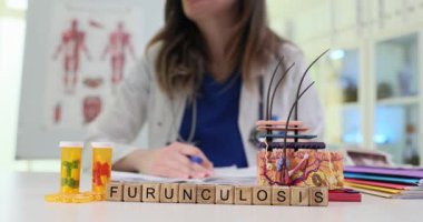 Masanın üzerinde kelime Furunulosis ile Scrabble taşları var. Kadın derisi modelinin yanında. Kadın doktor, modern klinikteki evrak işlerine odaklanmış.