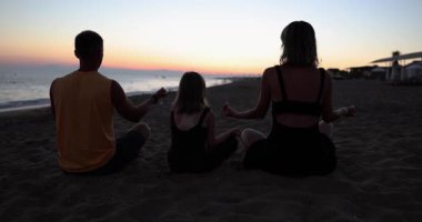 Küçük kızı olan bir aile, gün batımında sahilde yan yana oturup meditasyon yapıyor. Ebeveynler ve kızlar yaz tatilinde farkındalığın tadını çıkarırlar.