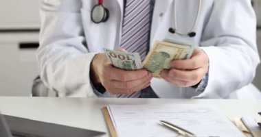 Beyaz cüppeli doktor klinikte oturmuş para banknotlarını sayıyor. Tıbbi uzman hasta randevusundan sonra nakit rüşveti kontrol ediyor.