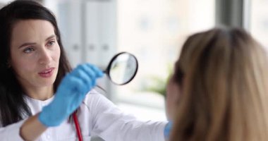Kadın dermatolog elinde büyüteç tutuyor ve kadın hastanın yanağındaki kızarıklıkları inceliyor. Doktor tahriş ya da enfeksiyon belirtilerine bakıyor.