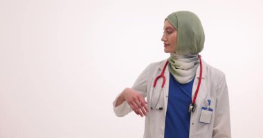Açık yeşil başörtüsü takan genç bir kadın doktor avuç içi ve işaret parmağıyla beyaz duvara yaslanmış. Tıbbi profesyonellik objeye dikkat çekiyor