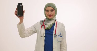 Müslüman kadın doktor, avuç içlerine vitamin dökerek siyah şişeyi açıyor. Steteskoplu tıbbi kıyafet giyen kadın, ağır çekimde gülümseyen harekete odaklanıyor.
