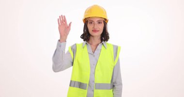 Sarı şapkalı ve yüksek görüşlü yelekli bir kadın selamlaşırken gülümsüyor. Kadın işçi, aydınlık odada duran mühendis meslektaşlarını selamladı