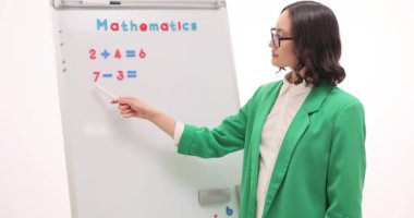 Kadın öğretmen matematik problemlerini beyaz tahtada açıklıyor. Kadın pedagog ilkokul derslerinde toplama ve çıkarma görevlerini yavaş çekimde gösterir