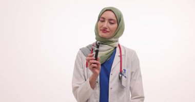 Düzgünce sarılmış tesettürdeki Otolaryngologist, beyaz arka plana karşı elde tutulan bir otoskop tutuyor. Doktor gülümser ve check-up için hazırlanır. Yavaş çekim