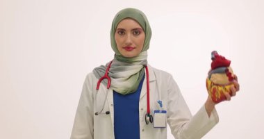 Türbanlı kadın kardiyolog beyaz arka planda insan kalbinin anatomik modelini tutuyor. Doktor kalp ilacı paketini kaldırıyor. Yavaş çekim