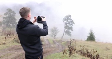 Siyah kapüşonlu ceketli bir adam doğada durup fotoğraf çekmek için kamera tutuyor. Etrafı sisli ve dağlık ağaçlarla çevrili.