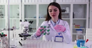 Laboratuvar önlüklü ve eldivenli genç bir kadın laboratuvarda moleküler modeli açıklıyor. Bayan, laboratuvar malzemeleriyle masada oturan yapıyı analiz ediyor.
