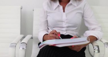 Beyaz gömlekli ve siyah etekli kadın sandalyeye oturmuş kalemle not defterine yazıyor. Ellere ve yazma eylemine odaklı ofis profesyonel ayarları