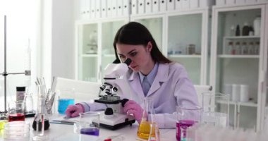Laboratuvar önlüğü ve pembe eldivenli kadın mikroskopla çalışıyor ve sonuçları laboratuarda yazıyor. Çeşitli laboratuvar ekipmanı ve reaksiyonlarıyla deney