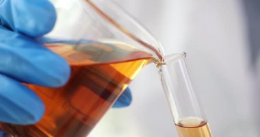 Mavi eldivenli bilim adamı konik şişeden test tüpüne sıvı döküyor. Kahverengi sıvı, laboratuvarda kimyasal ve bilimsel deney yapılmasını öneriyor.