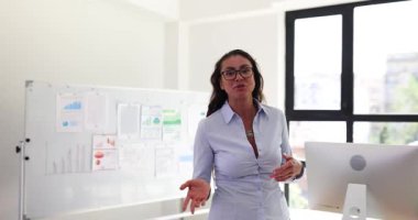 Gözlüklü kadın ofis ortamında yürürken elleri ve konuşmalarıyla el kol hareketi yapıyor. İşçi odadaki projeyi beyaz tahta ve bilgisayar monitörüyle sunar