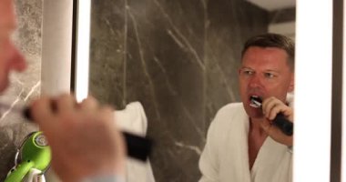 Bornozlu bir adam oteldeki modern banyo aynasının yanında elektrikli diş fırçasıyla dişlerini fırçalıyor. İnsan ağız sağlığını korumaya odaklanır.