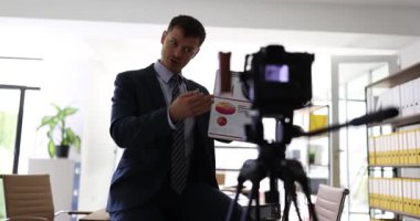 Ekonomi koçu masanın üstünde oturmuş eğitim finans videosu kaydediyor. Adam ofisindeki kameraya ticari süreçleri açıklayan grafiklere işaret ediyor.