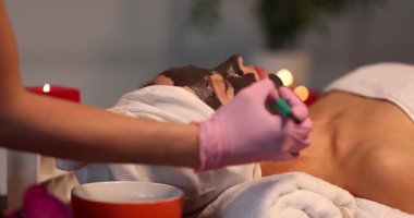 Pembe eldivenli güzellik uzmanı kuaförde genç bir kadının yüzüne siyah kilden maske takıyor. Dermatolog, kaplıca merkezindeki hasta cildiyle ilgilenir.